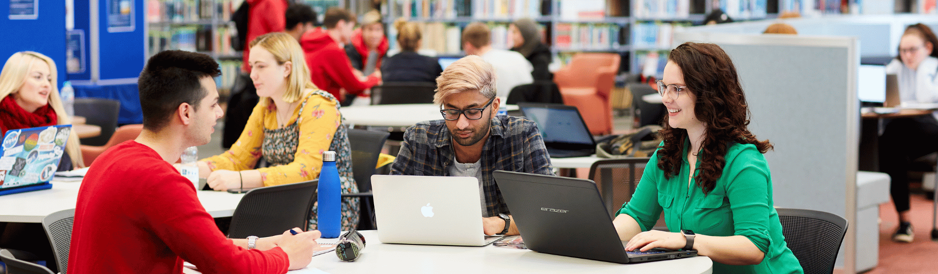 3 students sitting around a desk with their laptops open discussing work.