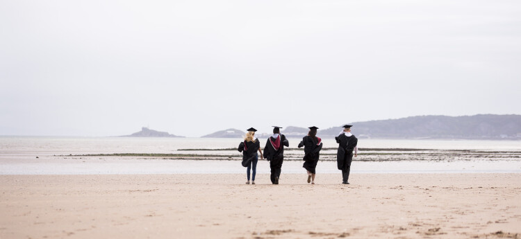 Four students walking on a beach in graduation gowns 