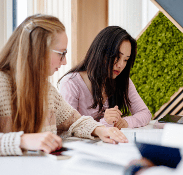 Two student sitting down and looking at a paper on a desk.